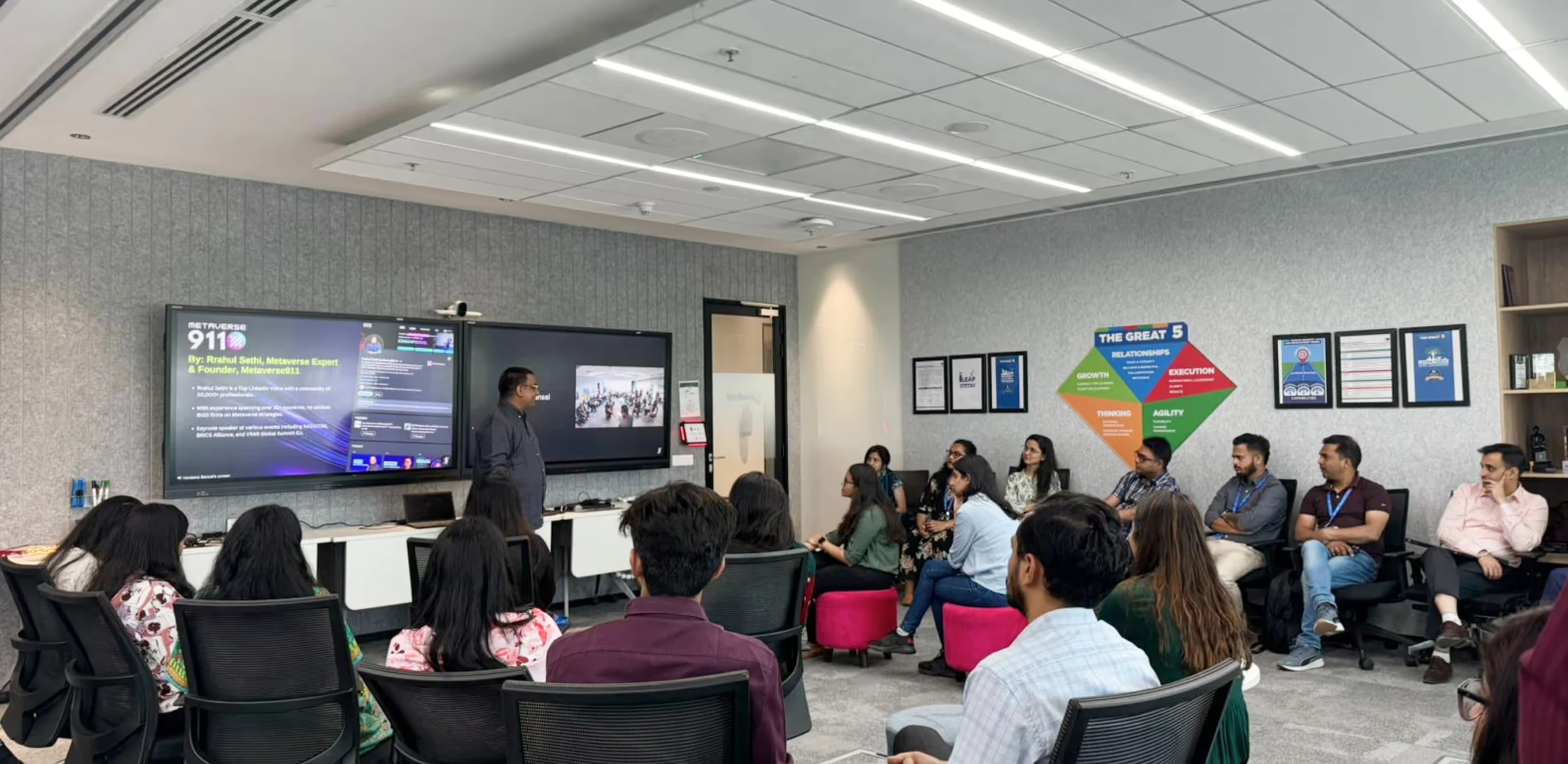 A man giving a presentation to a group of people seated in a conference room with two large screens displaying Metaverse 911 information and a video conference.