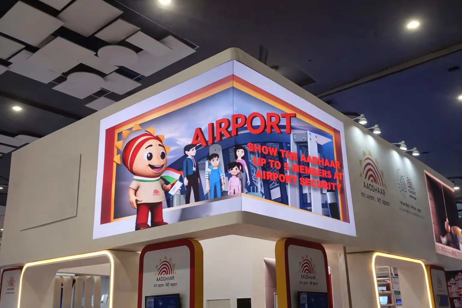 Digital display at an Aadhaar booth showing a cartoon character holding an Indian flag and text advising to show Aadhaar for up to five members at airport security.
