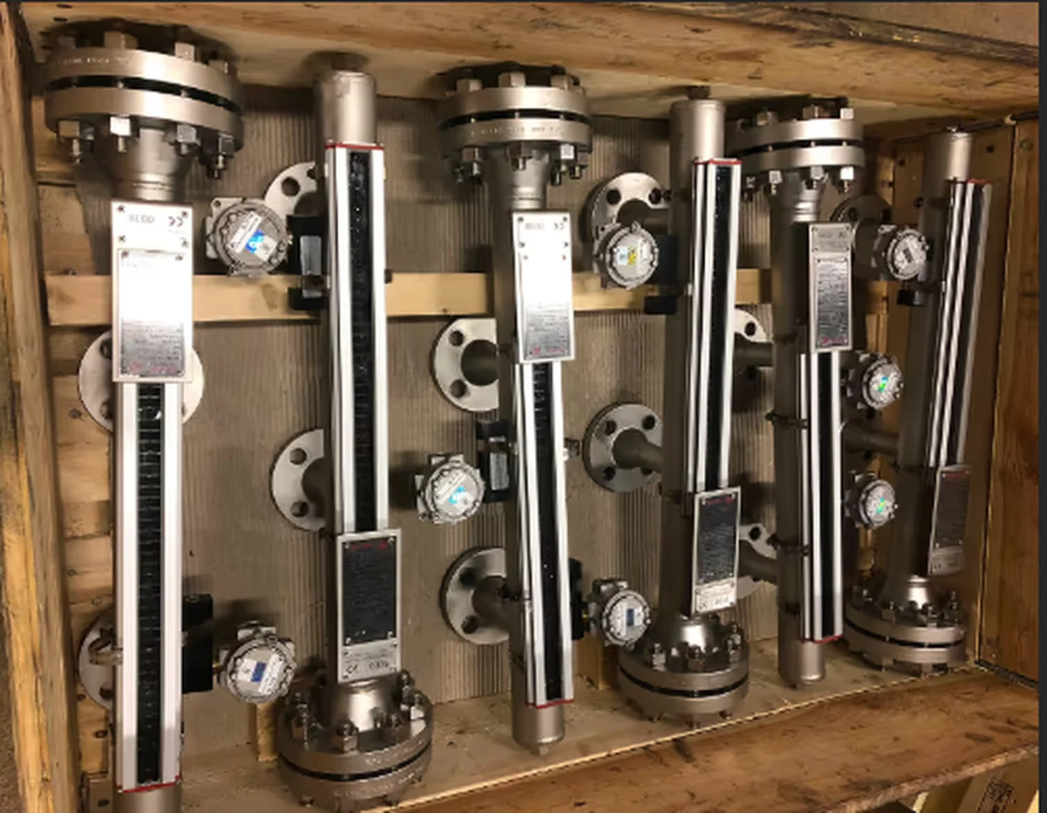 Five industrial stainless steel level gauge instruments with flange mounts arranged in a wooden crate.