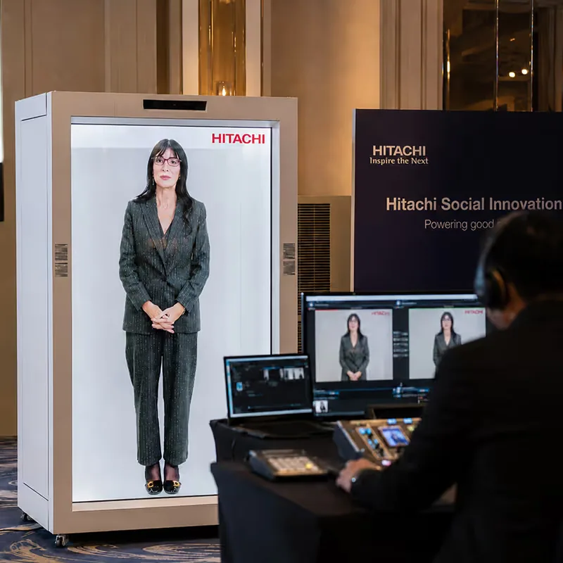 HoloBox911 unit on stage in front of a Hitachi Social Innovation Powering good backdrop, with a crew member alongside, at Hitachi's company event in New Delhi