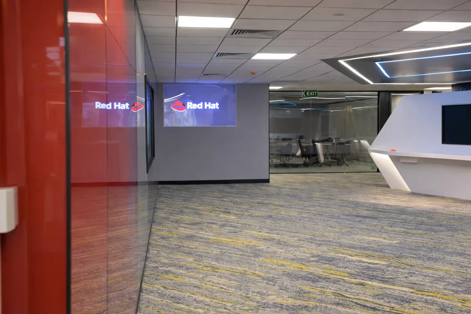 Modern office corridor with Red Hat logo displayed on a screen, glass-walled meeting room, and illuminated white reception desk.