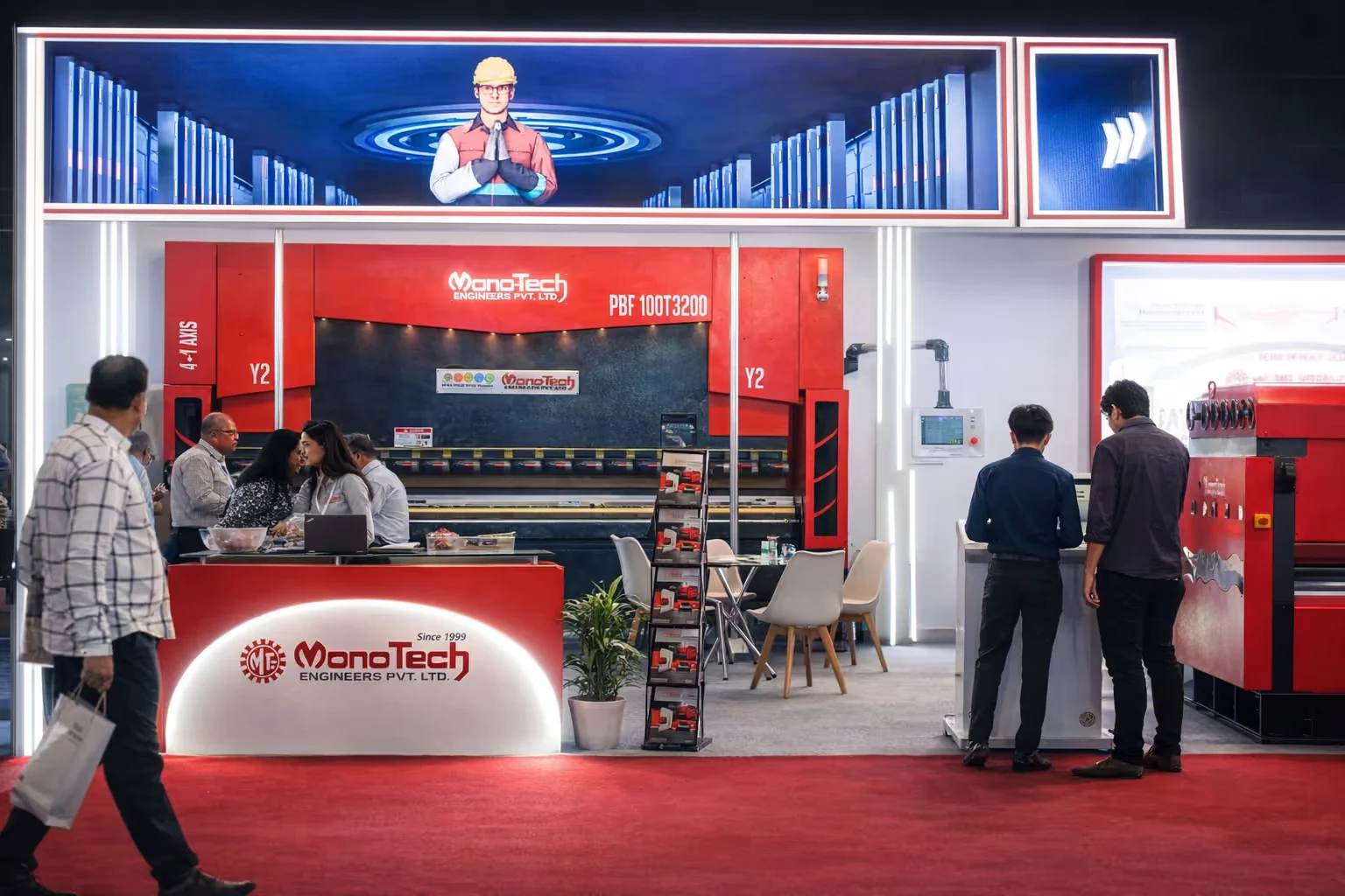 Exhibition booth of MonoTech Engineers Pvt. Ltd. displaying industrial machinery with people interacting and red carpet flooring.