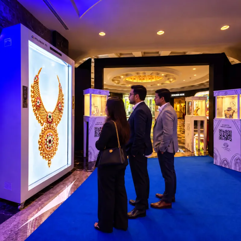 Visitors at a HoloBox911 display showing a gold necklace set beside exhibitor cabinets at COUTURE INDIA 2026, New Delhi