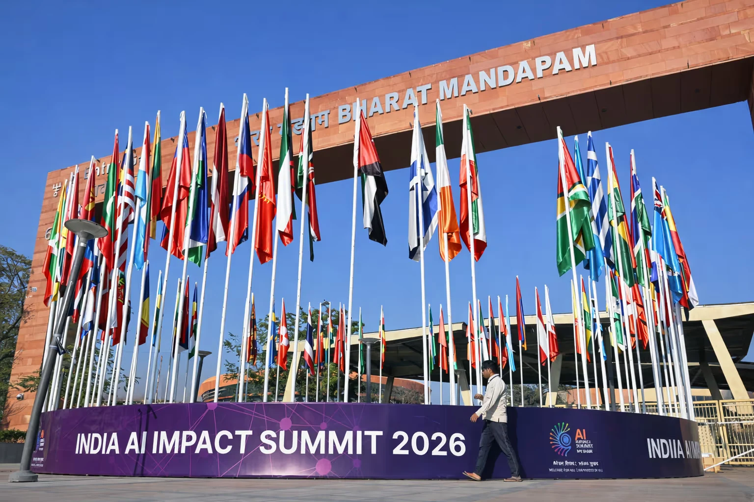 Circular display of multiple international flags in front of Bharat Mandapam with a banner reading India AI Impact Summit 2026.