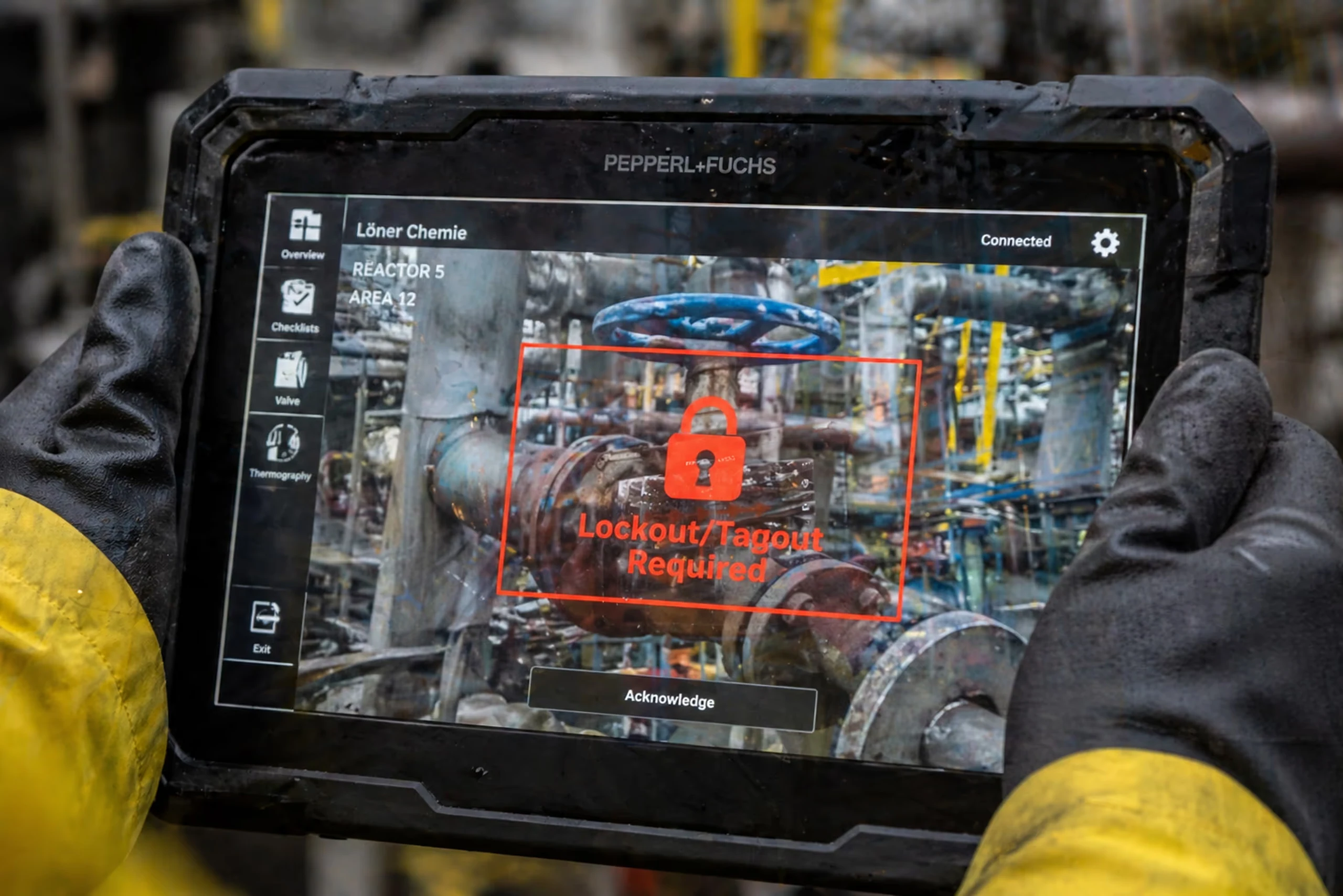 Industrial worker wearing gloves holding a rugged tablet displaying a red lock warning with 'Lockout/Tagout Required' over a valve in a chemical plant.