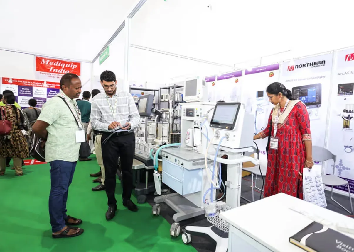 Exhibition booth photo of visitors examining a medical ventilator and diagnostic equipment at the Mediqip India stand, with a sales representative explaining product details on a green-carpeted trade show floor.