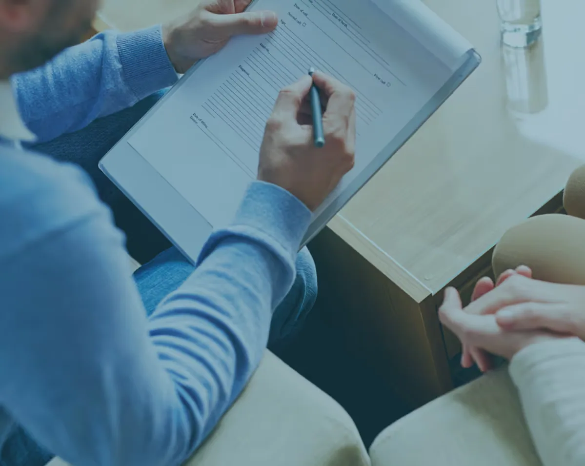 Person filling out a form on a clipboard while another person sits nearby with hands clasped.